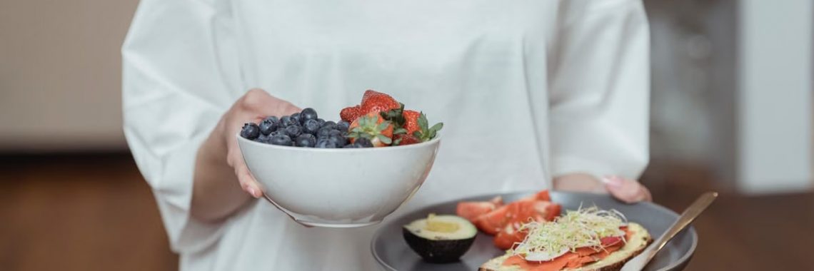 Person holding a bowl of fresh berries and a plate with avocado toast, displaying a healthy breakfast spread.